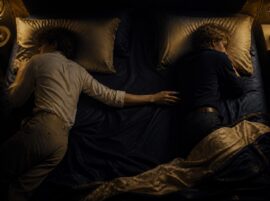 Two people resting in a softly lit bed with a small space between them, one hand reaching across the sheets, representing intimacy changes after menopause