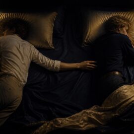 Two people resting in a softly lit bed with a small space between them, one hand reaching across the sheets, representing intimacy changes after menopause